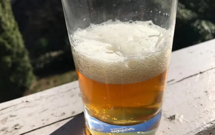 Ein Glas frisches, klares Bier mit einer schönen Schaumkrone steht auf einer weißen Holzoberfläche im Freien.